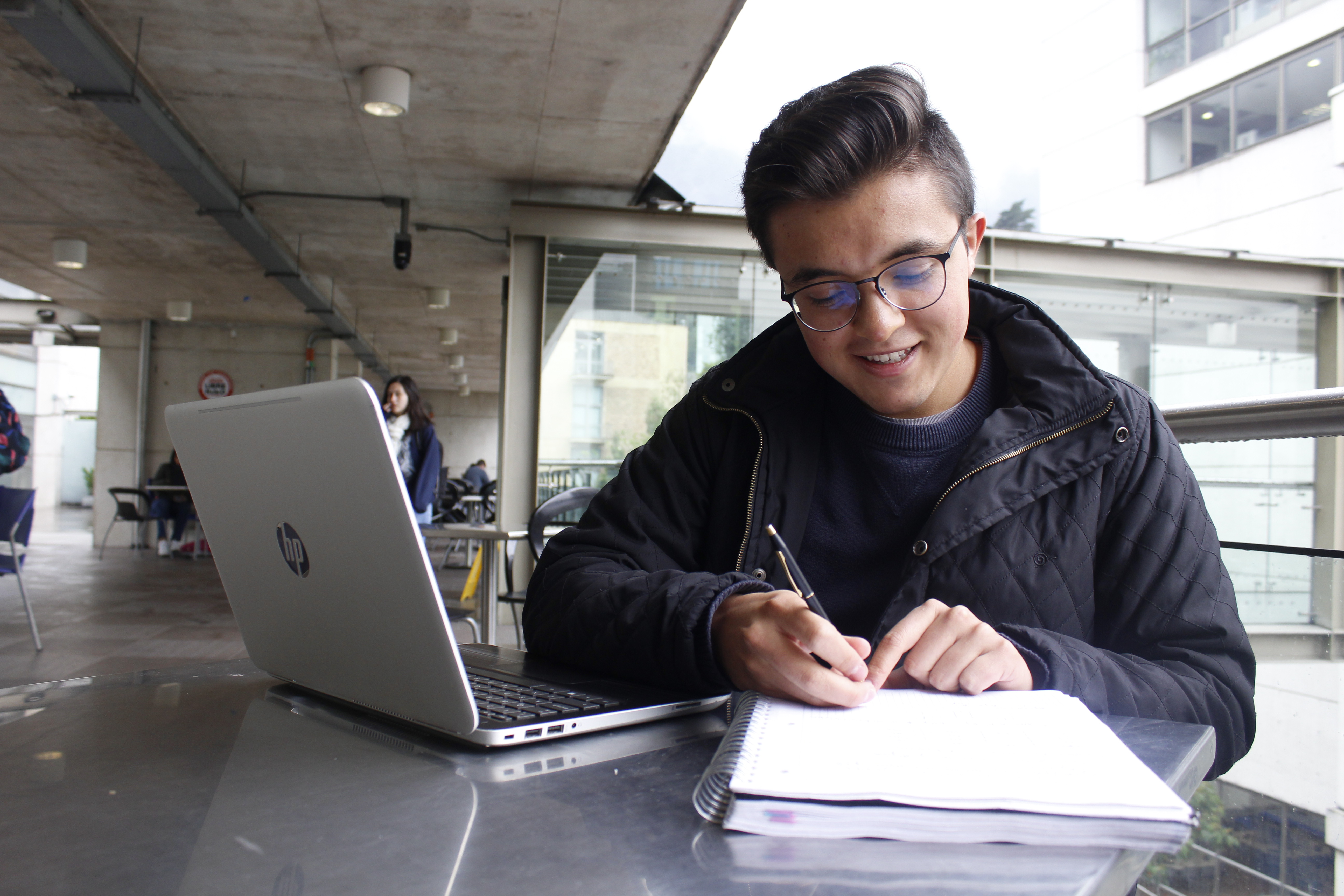 Estudiante tomando apuntes al aire libre mientras usa una computadora portátil.